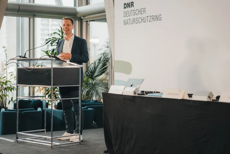Blick auf das Podium und ein Stehpult, Kai Niebert begrüßt die Gäste, im Hintergrund eine Leinwand mit Logo und Aufschrift "DNR Deutscher Naturschutzring"